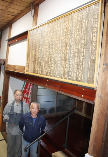 位牌堂入口に飾られた般若心経の木彫。岡村住職と上野さん(左から)=医王寺で