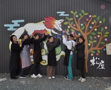 壁に描かれた絵の前で絵馬を持つスタッフと久保田さん(左端)=松屋で
