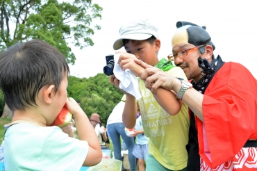 橘田さん㊨のアドバイスを聞きながらスイカを食べる幼児を撮影する少年=岩屋緑地で