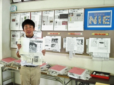 壁に貼られた「NIE通信」やスクラップブックの前に立つ金田教諭
