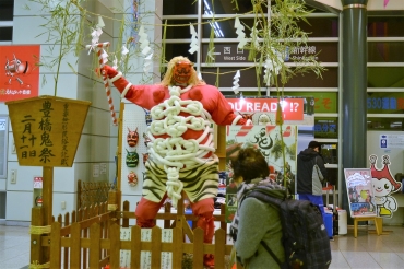 豊橋鬼祭をPRしているね赤鬼人形。後方は豊城中学生徒が作った張り子の面=豊橋駅東西自由連絡通路で