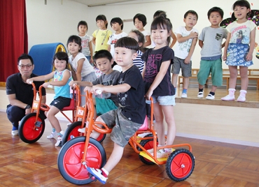 プレゼントされた三輪車に乗る園児と鈴木さん(左奥)=蒲郡市西浦保育園で